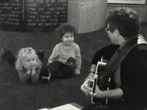 Photo of Lisa with guitar sitting on the Bema at Micah with a rapt Eden and Zach (toddlers) on the floor with her